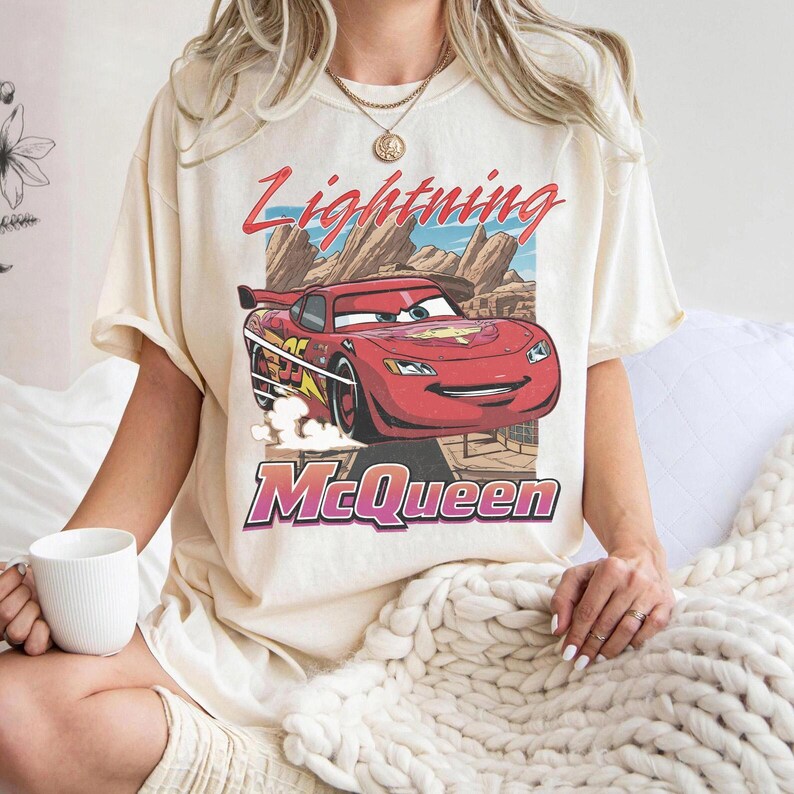 K&ouml;nnte beinhalten: Cremefarbenes T-Shirt mit einer Grafik von Lightning McQueen aus dem Film Cars. Das Design enth&auml;lt den Text "Lightning McQueen" in Rot und "McQueen" in Lila. Das Auto ist rot mit gelben und schwarzen Akzenten.