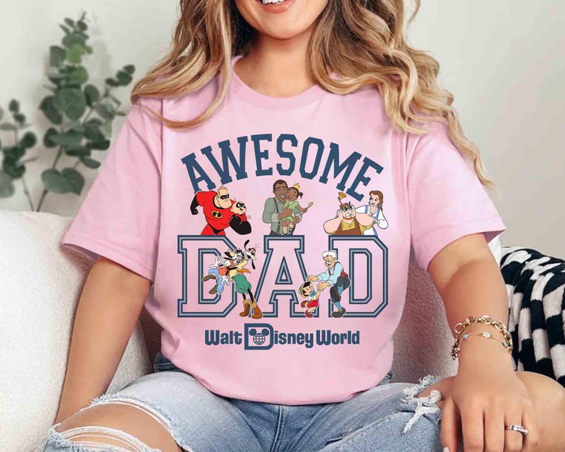 May include: Pink t-shirt with the words "AWESOME DAD" in blue block letters, featuring Disney characters. The shirt also includes the text "Walt Disney World" in blue. The design includes cartoon images of various Disney characters.