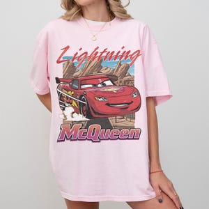 K&ouml;nnte beinhalten: Hellrosa T-Shirt mit einem Lightning McQueen-Motiv aus dem Film Cars. Das Design zeigt den Text "Lightning McQueen" in Rot und Lila, mit einem Cartoon-Bild des roten Rennwagens. Das T-Shirt hat kurze &Auml;rmel.