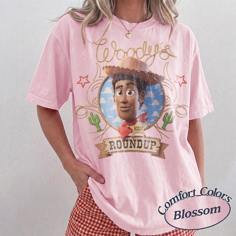 May include: A light pink short-sleeve t-shirt featuring a graphic of the character Woody from Toy Story inside a circular frame. The design includes the text Woody's Roundup, a lasso, stars, and cactus icons. The shirt has a crew neckline.