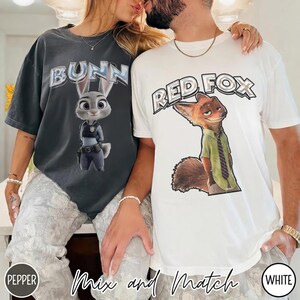 以下が含まれることがあります： 2枚のTシャツ。1枚は濃いグレー、もう1枚は白で、漫画のキャラクターが描かれています。濃いグレーのシャツには、警察官の制服を着たウサギの上に「Bunn」と書かれています。白いシャツには、緑のシャツとネクタイを着たキツネの上に「Red Fox」と書かれています。「Mix and Match」というフレーズが一番下にあります。