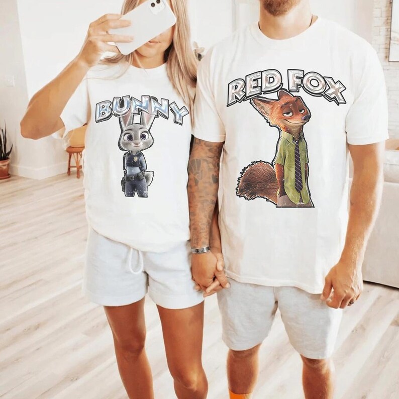 以下が含まれることがあります： 漫画のグラフィックがプリントされた白いTシャツ2枚。1枚は「Bunny」の文字とウサギのイラスト。もう1枚は「Red Fox」の文字とキツネのイラスト。どちらもライトグレーのショートパンツと合わせています。カップルの写真です。