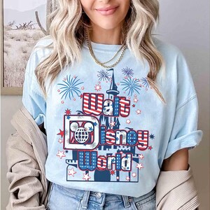 May include: Light blue t-shirt with a graphic featuring the text "Walt Disney World" in red, white, and blue. The design includes a castle, fireworks, and Mickey Mouse imagery. The shirt is a casual style.