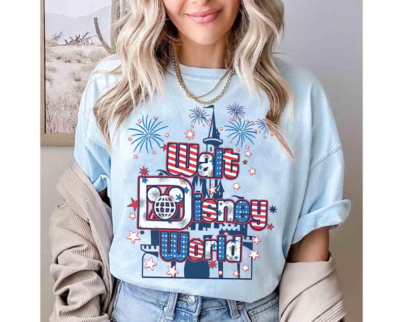 May include: Light blue t-shirt with a graphic featuring the text "Walt Disney World" in red, white, and blue. The design includes a castle, fireworks, and Mickey Mouse imagery. The shirt is a casual style.
