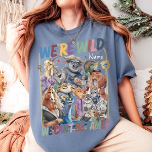 May include: A blue t-shirt featuring a colorful graphic of various cartoon animal characters. The text "WE'RE WILD" is at the top, with space for a name, and "WE CAN'T BE AMED" at the bottom. The design includes a badge.