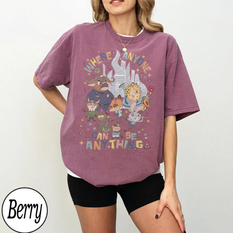 May include: Berry-colored t-shirt with a colorful graphic featuring cartoon characters and the text "WHERE ANYONE CAN BE ANYTHING". The shirt has a crew neck and short sleeves.