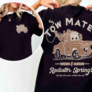 May include: A black t-shirt featuring a graphic of the character Tow Mater on the back with the text Tow Mater, Towing & Salvage, Radiator Springs, and It's like tuh-mater, without the tuh. A smaller version of the truck is printed on the front.