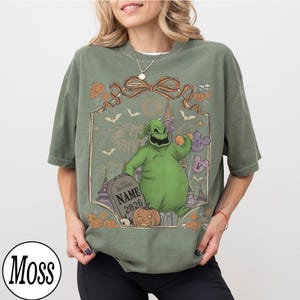K&ouml;nnte beinhalten: Olivgr&uuml;nes T-Shirt mit einem Halloween-Motiv. Das Design zeigt eine gr&uuml;ne Figur, K&uuml;rbisse, Flederm&auml;use, Feuerwerk und einen Grabstein mit dem Text "NAME 2026". Das T-Shirt hat eine lockere Passform und kurze &Auml;rmel.