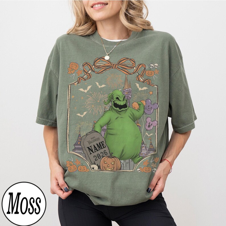 K&ouml;nnte beinhalten: Olivgr&uuml;nes T-Shirt mit einem Halloween-Motiv. Das Design zeigt eine gr&uuml;ne Figur, K&uuml;rbisse, Flederm&auml;use, Feuerwerk und einen Grabstein mit dem Text "NAME 2026". Das T-Shirt hat eine lockere Passform und kurze &Auml;rmel.