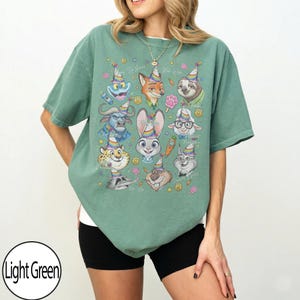 May include: Light green t-shirt featuring a colorful graphic of cartoon animal characters wearing party hats. The shirt has a relaxed fit and short sleeves. The text "Do Your Best, Be Wild" is also visible.