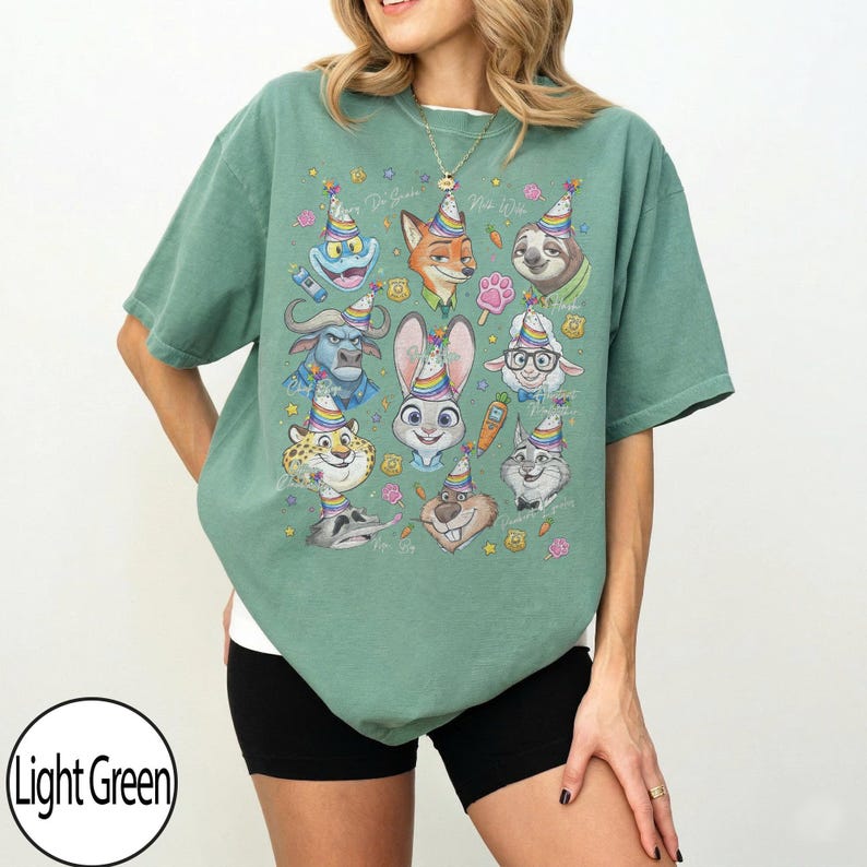 May include: Light green t-shirt featuring a colorful graphic of cartoon animal characters wearing party hats. The shirt has a relaxed fit and short sleeves. The text "Do Your Best, Be Wild" is also visible.