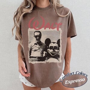 May include: Espresso-colored t-shirt featuring a vintage photo of Walt Disney with Mickey Mouse, both wearing sunglasses. The word "Walt" is printed above the photo in a pink, cursive font. The shirt has a Comfort Colors label.