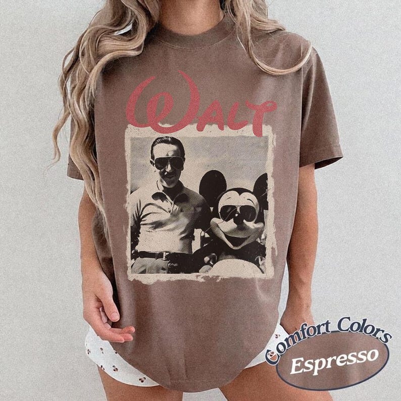 May include: Espresso-colored t-shirt featuring a vintage photo of Walt Disney with Mickey Mouse, both wearing sunglasses. The word "Walt" is printed above the photo in a pink, cursive font. The shirt has a Comfort Colors label.
