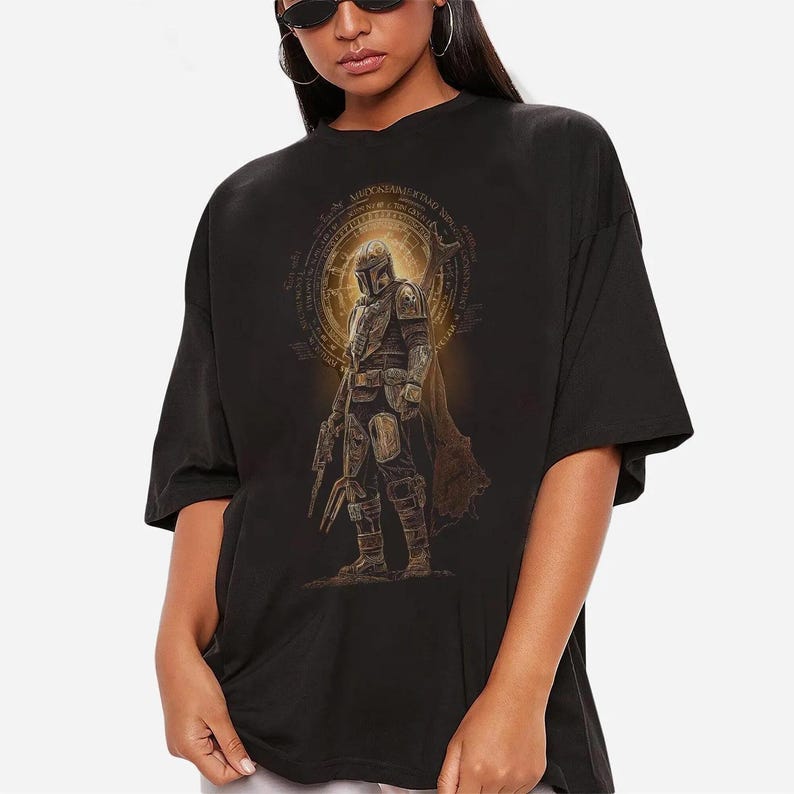 May include: A black oversized t-shirt featuring a graphic print of a armored character in a helmet and cape, standing in front of a glowing golden circular emblem with intricate text details. The design is centered on the chest of the garment.