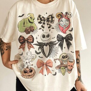 May include: An off-white t-shirt featuring a graphic print of characters and bows from The Nightmare Before Christmas. The design includes Jack Skellington, Sally, Oogie Boogie, Zero, and Lock, Shock, and Barrel, interspersed with spiderwebs and bats.