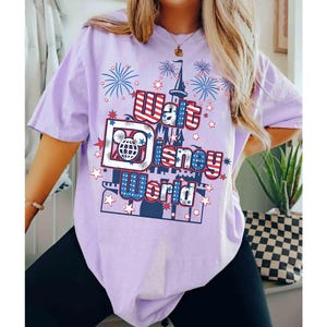 May include: A light purple t-shirt featuring a graphic design with the text "Walt Disney World" in red, white, and blue. The design includes a castle, fireworks, and stars. The shirt is a short-sleeved, casual style.