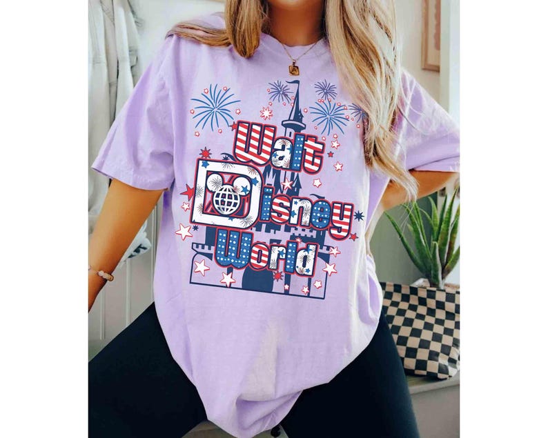 May include: A light purple t-shirt featuring a graphic design with the text "Walt Disney World" in red, white, and blue. The design includes a castle, fireworks, and stars. The shirt is a short-sleeved, casual style.