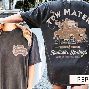 May include: A charcoal gray t-shirt featuring a graphic of the character Tow Mater on the back with the text Tow Mater, Towing & Salvage, Radiator Springs, and It's like tah-mater, without the tah. A smaller version of the truck is on the front.