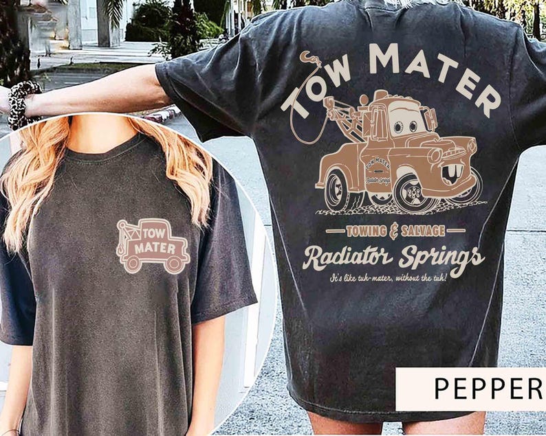May include: A charcoal gray t-shirt featuring a graphic of the character Tow Mater on the back with the text Tow Mater, Towing & Salvage, Radiator Springs, and It's like tah-mater, without the tah. A smaller version of the truck is on the front.