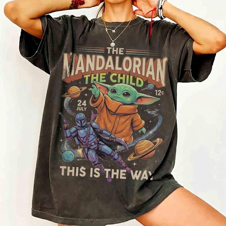May include: A charcoal gray short-sleeve t-shirt featuring a graphic print of The Mandalorian and The Child. The design includes space elements, planets, and the text "THE MANDALORIAN", "THE CHILD", "24 JULY", "12c", and "THIS IS THE WAY" in a vintage style.