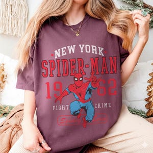 May include: A maroon t-shirt featuring a graphic of Spider-Man in a dynamic pose. The design includes the text "NEW YORK SPIDER-MAN 1962 FIGHT CRIME" in red and white. The shirt is a casual wear item.