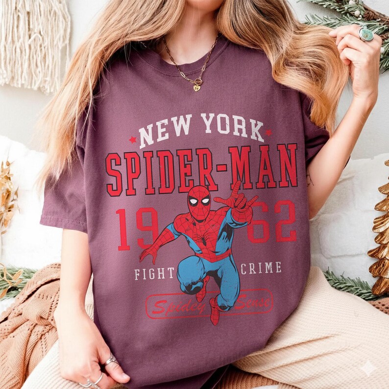 May include: A maroon t-shirt featuring a graphic of Spider-Man in a dynamic pose. The design includes the text "NEW YORK SPIDER-MAN 1962 FIGHT CRIME" in red and white. The shirt is a casual wear item.