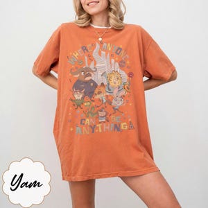 May include: Orange t-shirt featuring a colorful graphic of animated characters and the text "WHERE ANYONE CAN BE ANYTHING". The design includes a castle and various animal figures. A casual, short-sleeved tee.