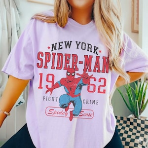 May include: A light purple t-shirt featuring a graphic of Spider-Man. The design includes the text "NEW YORK SPIDER-MAN 1962 FIGHT CRIME" in red, with an illustration of Spider-Man in his classic red and blue suit. The words "Spidey Sense" are also included.