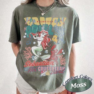 May include: Moss green t-shirt featuring a graphic of Ariel from The Little Mermaid, with text that reads "A LANTICA HOTEL OCEAN TOUR 1991" and "Sebastian's Hot Crustacean Band". The shirt has a vintage, distressed look.