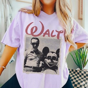 May include: A light purple t-shirt featuring a vintage-style image of Walt Disney with Mickey Mouse. The word "Walt" is printed above the image in a reddish-brown, textured font. The shirt is a casual style.