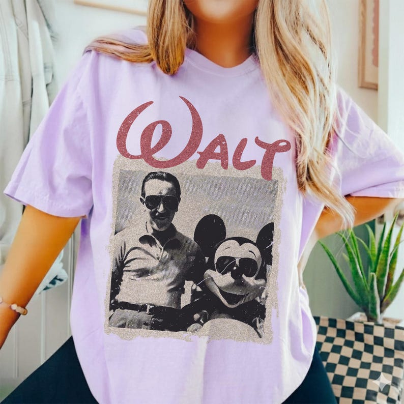 May include: A light purple t-shirt featuring a vintage-style image of Walt Disney with Mickey Mouse. The word "Walt" is printed above the image in a reddish-brown, textured font. The shirt is a casual style.