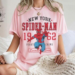May include: Pink t-shirt featuring a Spider-Man graphic. The design includes the text "New York Spider-Man 1962" in red, with an illustration of Spider-Man in his classic red and blue suit. The words "Fight Crime" and "Spidey Sense" are also included.