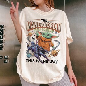 May include: A cream-colored t-shirt featuring a graphic print of The Mandalorian and The Child. The design includes planets, stars, and text that reads THE MANDALORIAN THE CHILD, 24 JULY, 12c, and THIS IS THE WAY in a vintage comic book style.