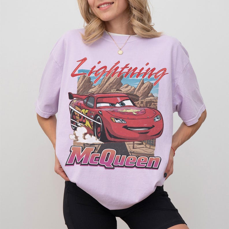 K&ouml;nnte beinhalten: Ein helllila T-Shirt mit einer Grafik von Lightning McQueen aus dem Film Cars. Das Design enth&auml;lt den Text "Lightning McQueen" in Rot und den Namen der Figur in einer lila Schrift. Der Hintergrund zeigt eine W&uuml;stenlandschaft.