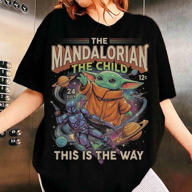 May include: A black t-shirt featuring a graphic print of The Mandalorian and The Child in space. The design includes planets, stars, and the text THE MANDALORIAN THE CHILD 24 JULY 12c THIS IS THE WAY. The characters are depicted in a colorful, retro style.