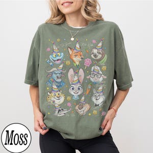May include: An olive green t-shirt featuring a colorful graphic of various cartoon animal characters wearing party hats. The design includes a frog, fox, sloth, bunny, and others. The shirt has the word "Moss" in a circle at the bottom.