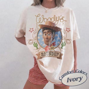 May include: An ivory t-shirt featuring a graphic of the character Woody from Toy Story inside a circular frame. The design includes the text Woody's and ROUNDUP, along with illustrations of a cowboy hat, stars, cacti, and a sheriff badge.
