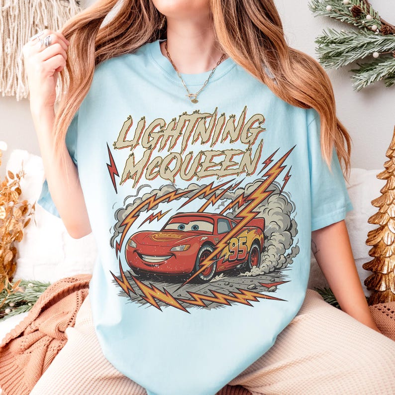 May include: Light blue t-shirt featuring Lightning McQueen from the Cars movie. The graphic shows the red race car with the number 95, surrounded by lightning bolts and smoke. The text "Lightning McQueen" is above the car.