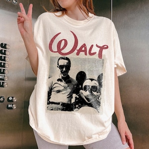 May include: Cream-colored t-shirt featuring a vintage-style graphic of Walt Disney and Mickey Mouse wearing sunglasses. The word "Walt" is printed in a distressed, burgundy font above the image. The shirt has a relaxed fit.