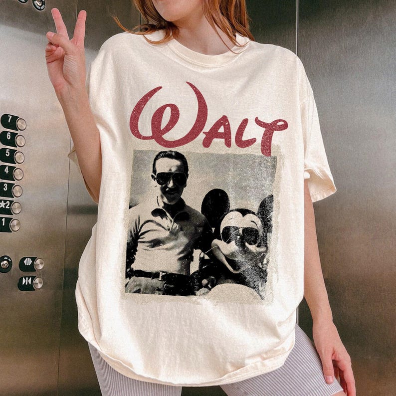May include: Cream-colored t-shirt featuring a vintage-style graphic of Walt Disney and Mickey Mouse wearing sunglasses. The word "Walt" is printed in a distressed, burgundy font above the image. The shirt has a relaxed fit.