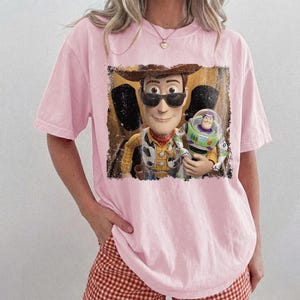 Puede incluir: Camiseta rosa claro con un gr&aacute;fico de Woody y Buzz Lightyear de Toy Story. Woody lleva gafas de sol y un sombrero de vaquero. Buzz Lightyear est&aacute; en su traje espacial. El gr&aacute;fico tiene un aspecto vintage.