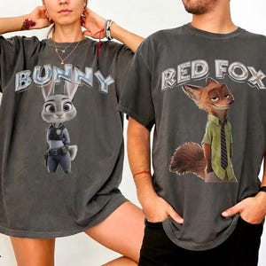 以下が含まれることがあります： ズートピアのキャラクターが描かれた2枚の濃いグレーのTシャツ。1枚のシャツには、警察の制服を着た漫画のウサギの上に「BUNNY」と表示されています。もう1枚のシャツには、緑のシャツとネクタイを着た漫画のキツネの上に「RED FOX」と表示されています。