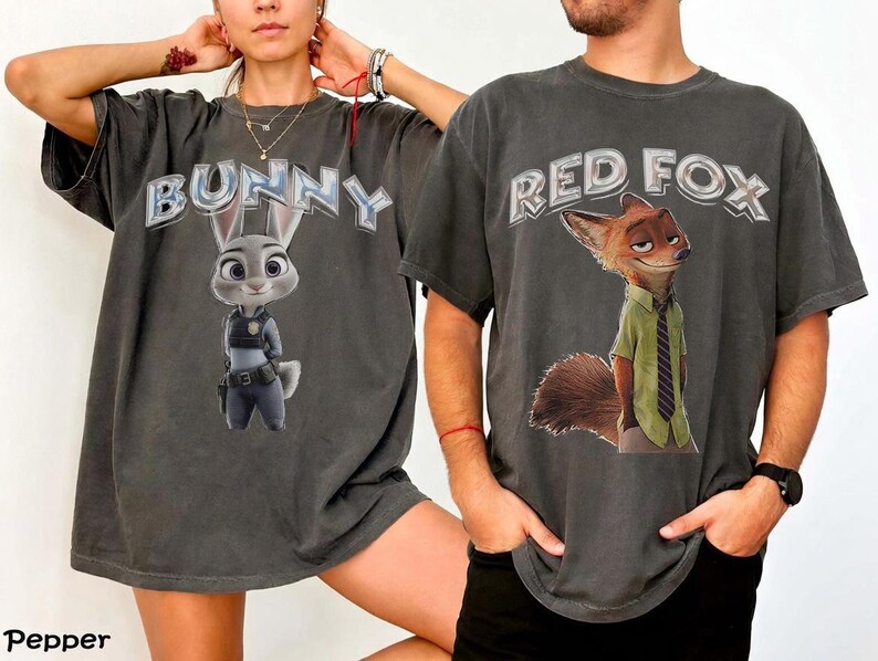 以下が含まれることがあります： ズートピアのキャラクターが描かれた2枚の濃いグレーのTシャツ。1枚のシャツには、警察の制服を着た漫画のウサギの上に「BUNNY」と表示されています。もう1枚のシャツには、緑のシャツとネクタイを着た漫画のキツネの上に「RED FOX」と表示されています。