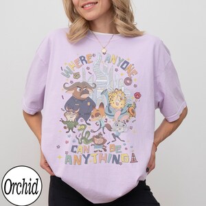 May include: A light purple t-shirt featuring cartoon characters and the text "WHERE ANYONE CAN BE ANYTHING". The design includes various animal characters in a colorful, whimsical style. The shirt has short sleeves and a relaxed fit.