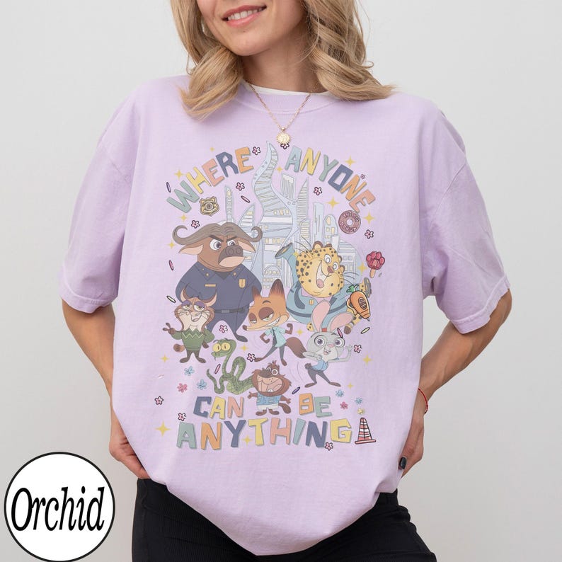 May include: A light purple t-shirt featuring cartoon characters and the text "WHERE ANYONE CAN BE ANYTHING". The design includes various animal characters in a colorful, whimsical style. The shirt has short sleeves and a relaxed fit.
