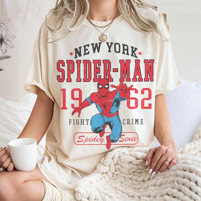 May include: Cream-colored t-shirt featuring a graphic of Spider-Man in red and blue, with the text "New York Spider-Man 1962 Fight Crime" in red and white. The shirt has a vintage, distressed look.