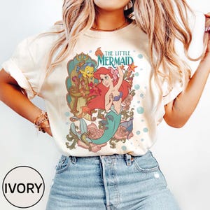 Pu&ograve; includere: T-shirt color avorio con una grafica colorata di Ariel de La Sirenetta. Il design include il testo "THE LITTLE MERMAID" in verde acqua, insieme a illustrazioni di Ariel, Flounder ed elementi floreali. La maglietta &egrave; abbinata a pantaloncini di jeans blu.