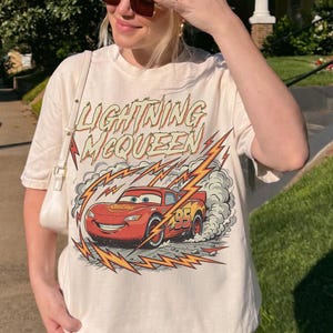 May include: Off-white t-shirt featuring a graphic of Lightning McQueen from the Cars movie. The design includes the text "Lightning McQueen" above the car and lightning bolt graphics. The number "95" is visible on the car.