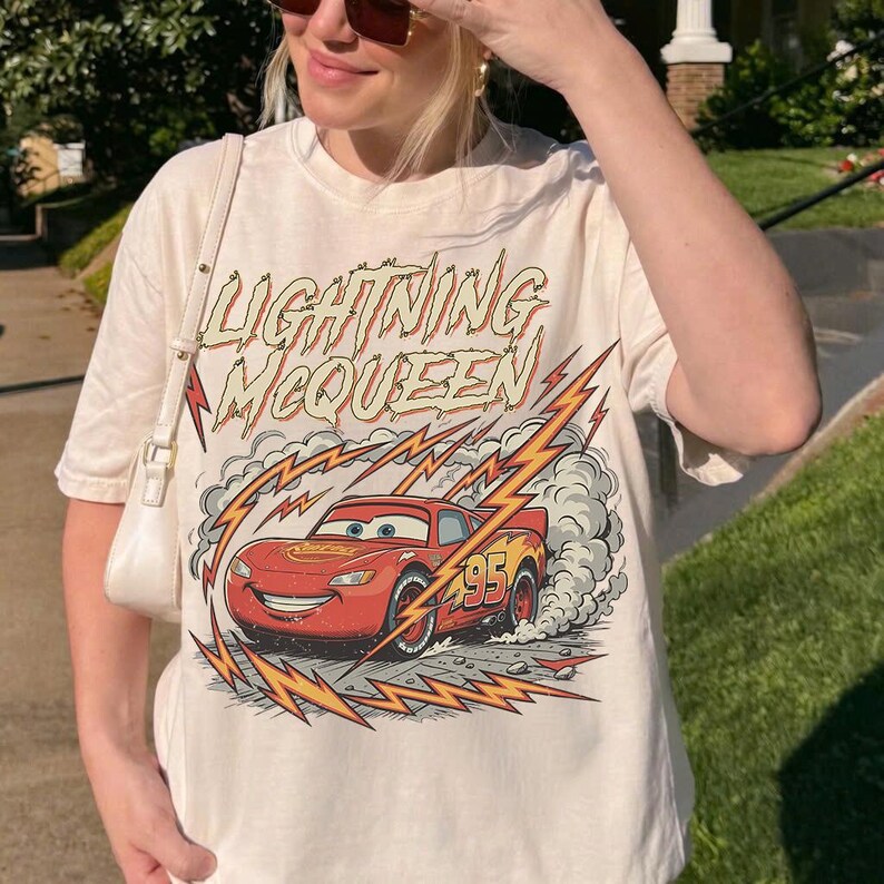 May include: Off-white t-shirt featuring a graphic of Lightning McQueen from the Cars movie. The design includes the text "Lightning McQueen" above the car and lightning bolt graphics. The number "95" is visible on the car.