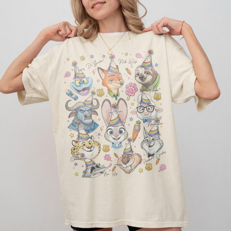 May include: Cream-colored t-shirt featuring colorful cartoon characters wearing party hats. The design includes a variety of animal characters with text, stars, and floral accents. The shirt is a relaxed fit.